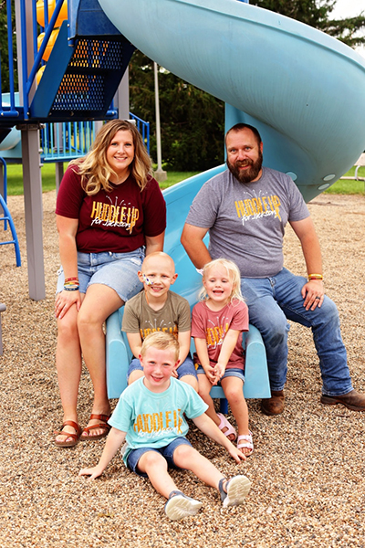 Family of five smiling together at a playground, all wearing matching “Huddle Up for Jackson” fundraiser t-shirts in different colors. Image represents community support and family strength through CAUSETEAM medical fundraising.